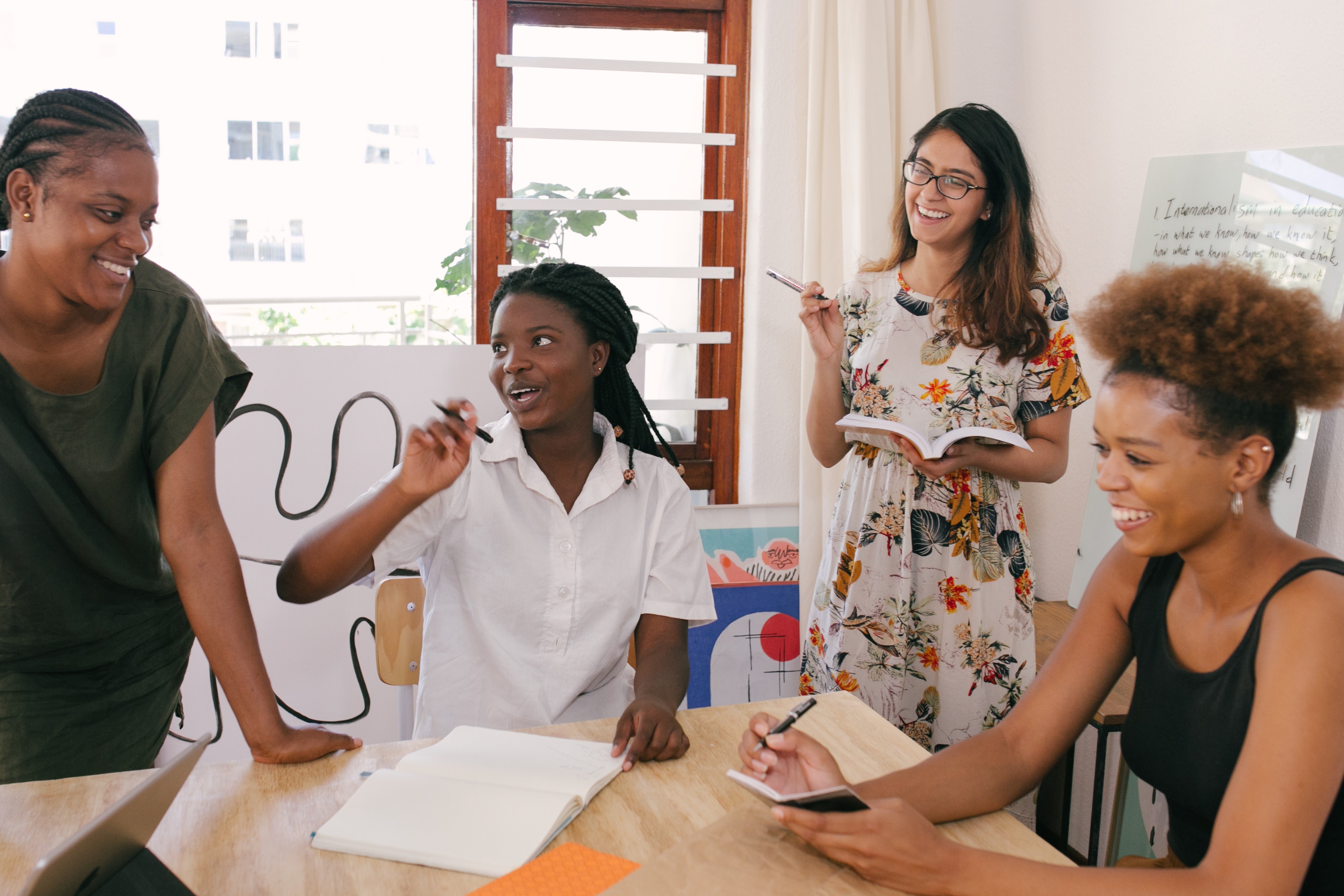 four young women of various races smile while working together in a sun-filled room