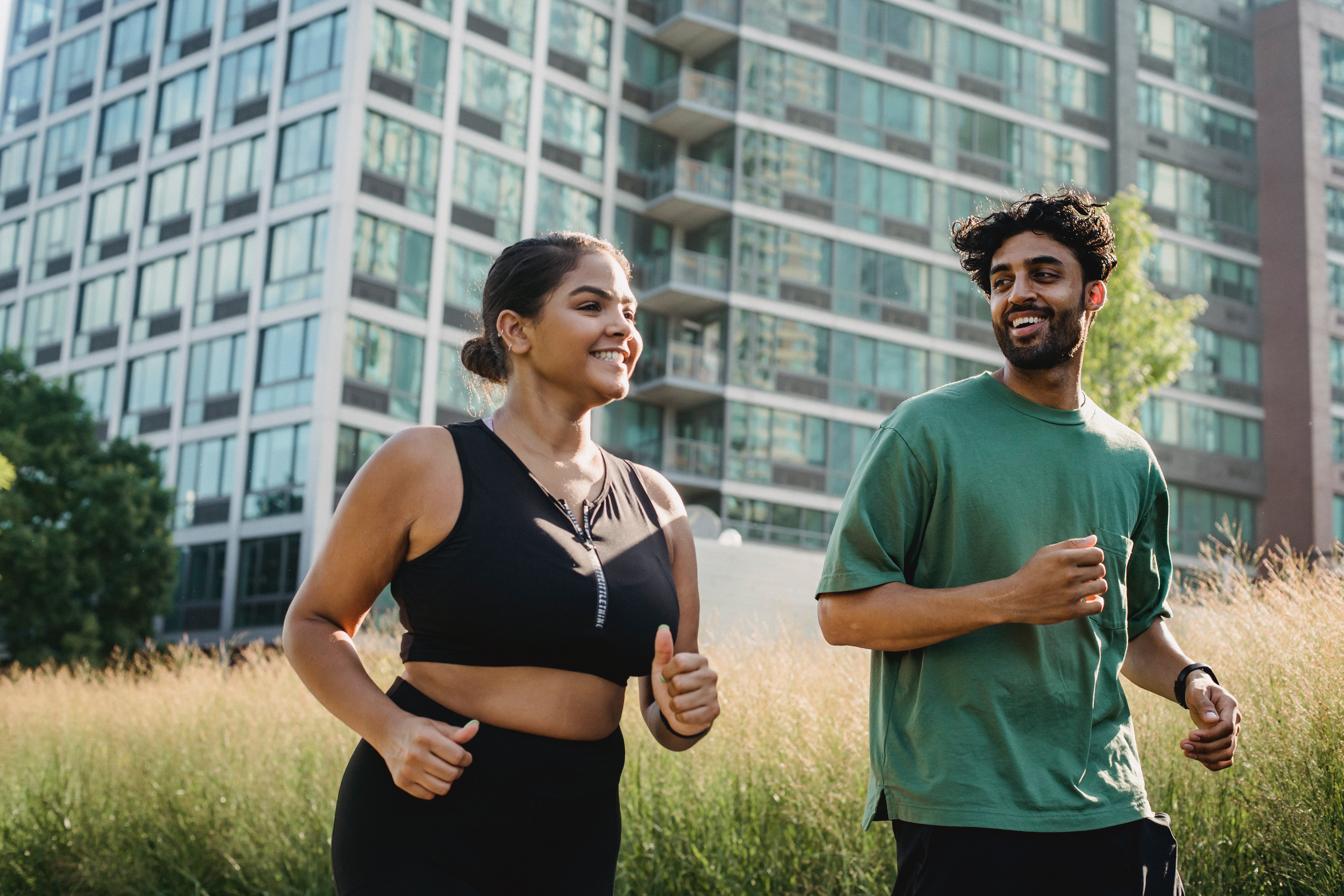 a young woman in a black tank top and a young man in a green shirt smile while they run outside in front of a large apartment building