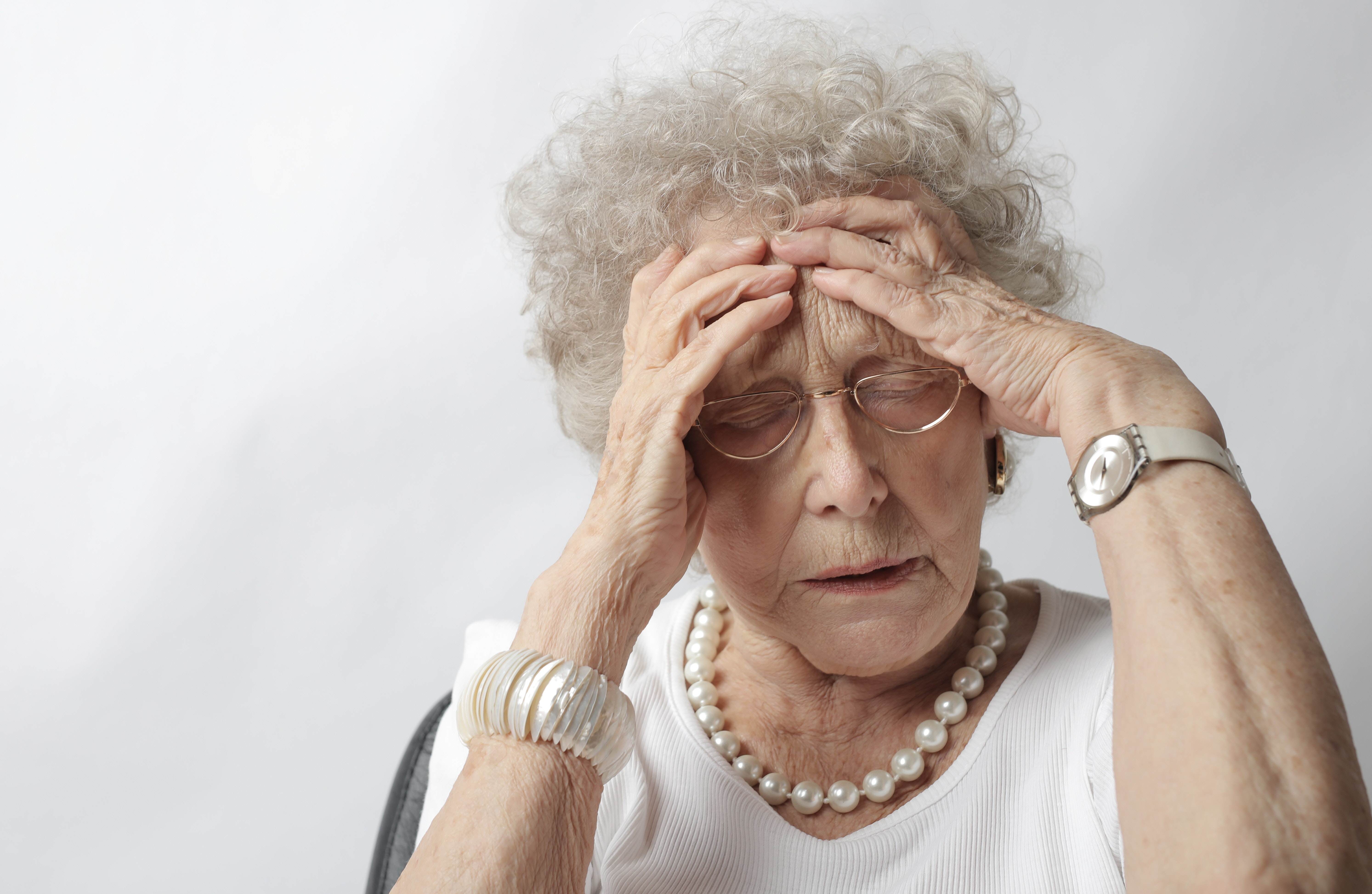 an older woman with white curly hair, wearing a white t-shirt and a pearl necklace, clutches at her forehead with both hands in distress